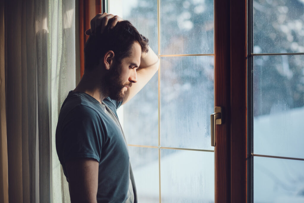 Man standing by window head down feeling fear and anxiety